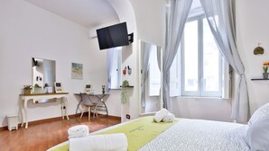 A hotel room with a bed, a desk, a chair, a TV, and a window with curtains. - GuestHost - Porta Medina Apartment (Napoli)