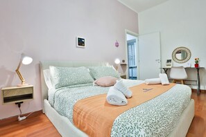 A neatly made bed with a patterned bedspread, a bedside lamp, a small bedside table, and a chair with a mirror.