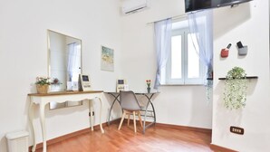 A room with a wooden floor, a white desk with a mirror, a chair, a small stool, a window with a curtain, and a wall-mounted shelf with a plant. - GuestHost - Porta Medina Apartment (Napoli)