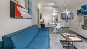 Una sala moderna con un sofá azul, un comedor con sillas blancas, una mesa con libros y un cuadro abstracto grande en la pared.