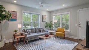 A living room with a sofa, armchair, coffee table, and a TV. There are large windows, a ceiling fan, and a door.