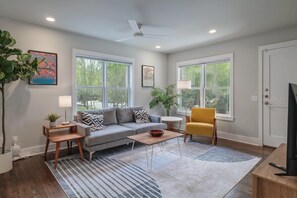 A living room with a sofa, armchair, coffee table, and a TV. There are large windows, a ceiling fan, and a door. - Quintessential Zilker Bungalow (Austin)