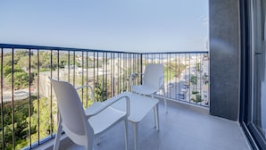 Un balcón con dos sillas blancas y una mesa, con vistas a un paisaje urbano con edificios y zonas verdes.