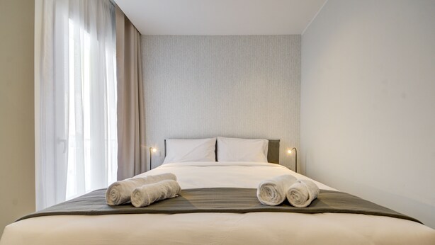 Een opgemaakt bed met twee opgerolde handdoeken, een eenvoudig hoofdbord en een lichtgekleurde muur.