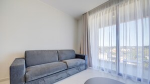 Een moderne woonkamer met een grijze bank, een ronde salontafel en lichte gordijnen voor een groot raam.