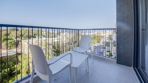 Un balcone con due sedie bianche e un tavolo, affaccia su un paesaggio urbano con edifici e vegetazione.