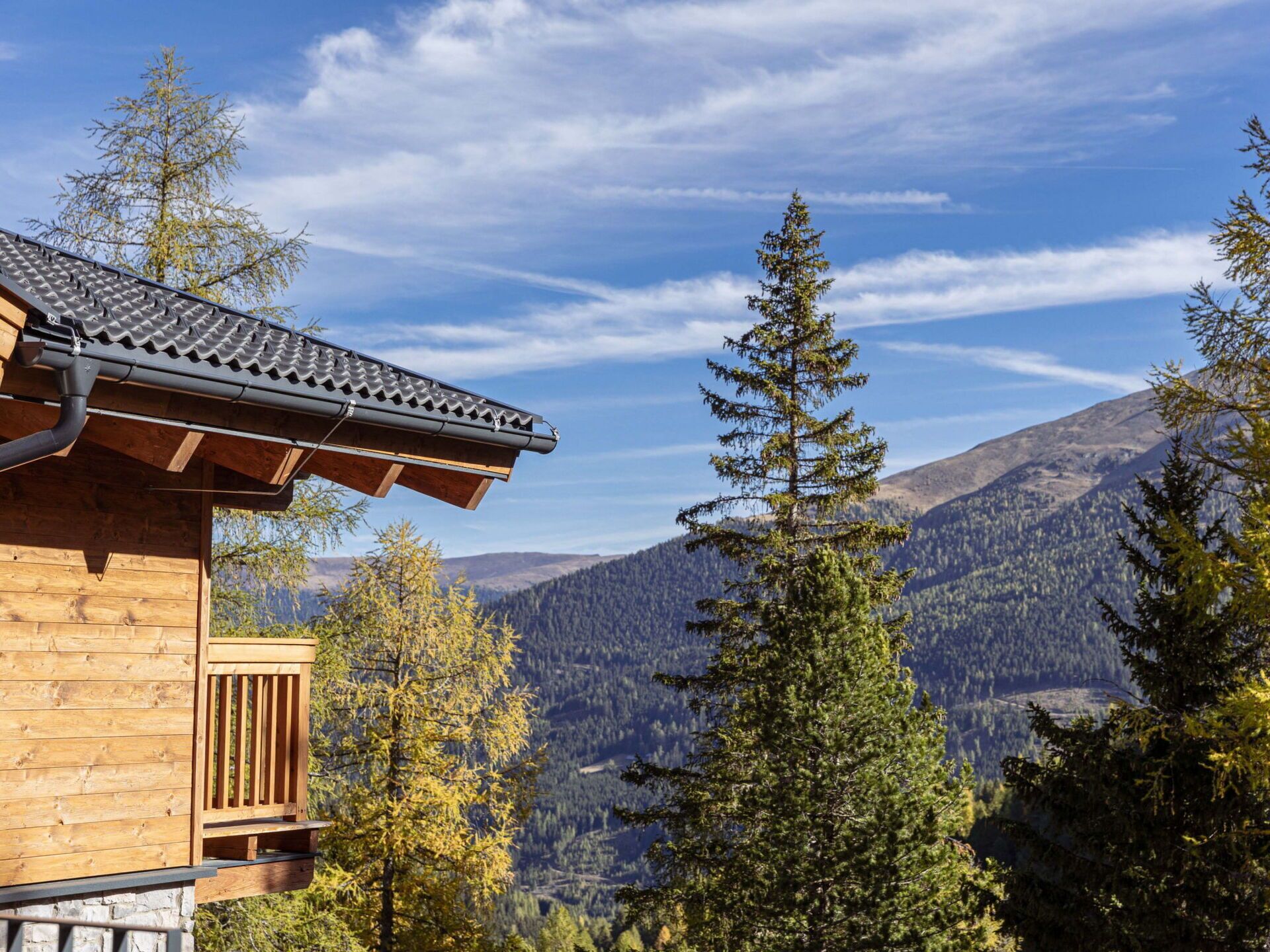 A wooden cabin with a tiled roof, surrounded by tall evergreen trees and a mountainous backdrop.
