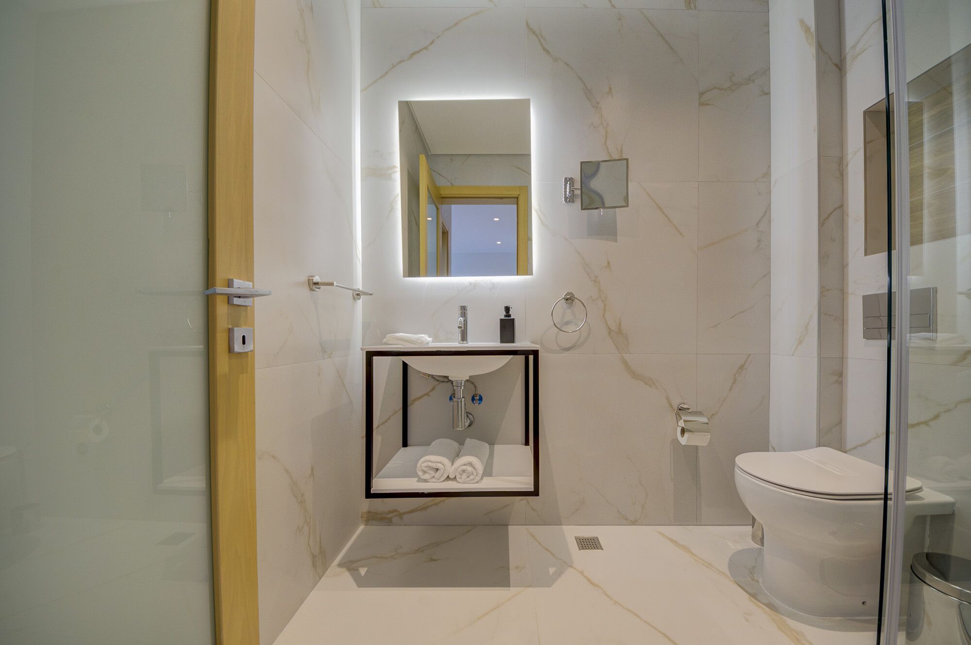 A modern bathroom with a glass shower enclosure, a marble-tiled floor, a built-in sink with a mirror, and a toilet.