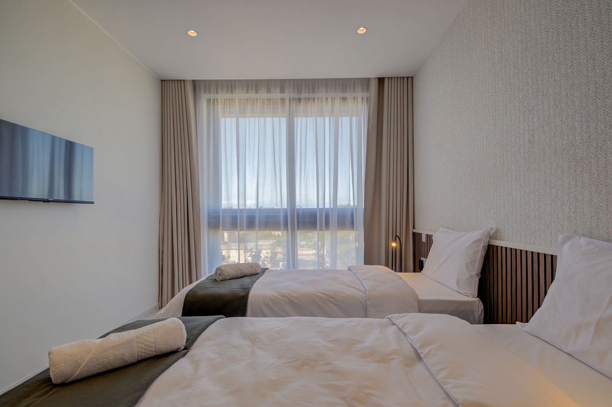 A hotel room with two beds, a large window with sheer curtains, a flat-screen TV mounted on the wall, and a textured wall.