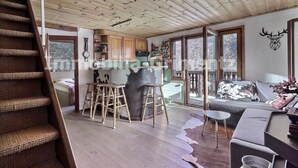 Ein gemütliches Cabin-Interieur mit einer Holztreppe, einer Küchenzeile mit Holzausstattung, einem Barhocker und einem großen Fenster mit Blick auf die verschneite Landschaft.