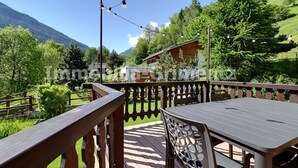 Eine Holzdauerdecke mit Tisch und Stühlen, mit Blick auf eine gebirgige Landschaft, im Hintergrund ein Holzhaus.