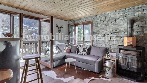 Ein gemütliches Blockhaus-Interieur mit einer Steinwand, einem grauen Sofa, einer Holzleiter und einem Kamin.