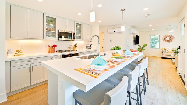 Una cocina moderna con armarios blancos, una isla central con taburetes y un área de comedor con sofá y televisor.