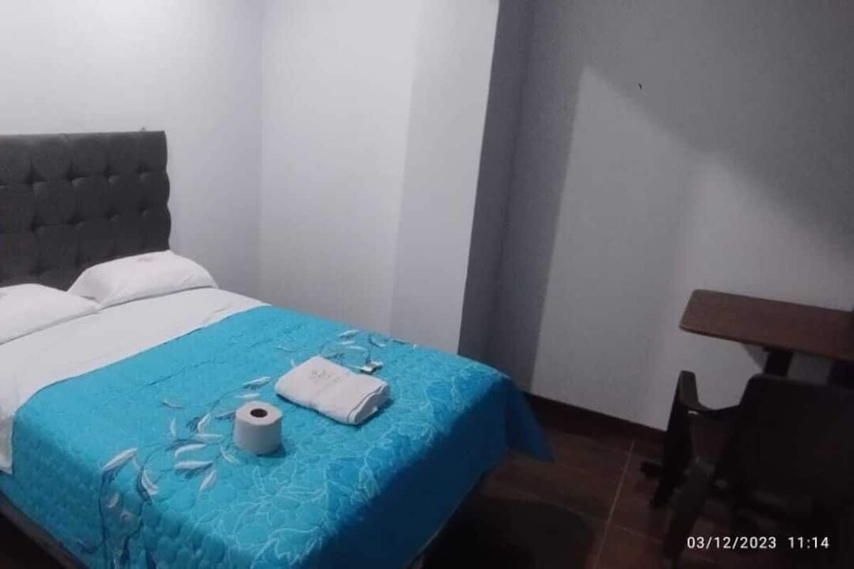 Ein Einzelbett mit grau gepolstertem Kopfteil, einer blauen Bettdecke und einem weißen Kissen. Ein Holznachttisch mit Schublade, eine Toilettenpapierrolle und ein gefaltetes Handtuch liegen auf dem Bett.