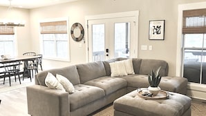 Una sala de estar moderna con un sofá modular, un comedor con mesa y sillas, y amplias ventanas con persianas.