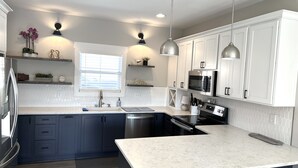 Una cocina moderna con armarios azul oscuro, encimeras blancas y electrodomésticos de acero inoxidable. Hay luces colgantes sobre la isla y una ventana con persianas.