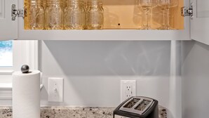 Una cocina con un mueble de madera que contiene frascos de vidrio y copas de vino, un tostador y un dispensador de toallas de papel.