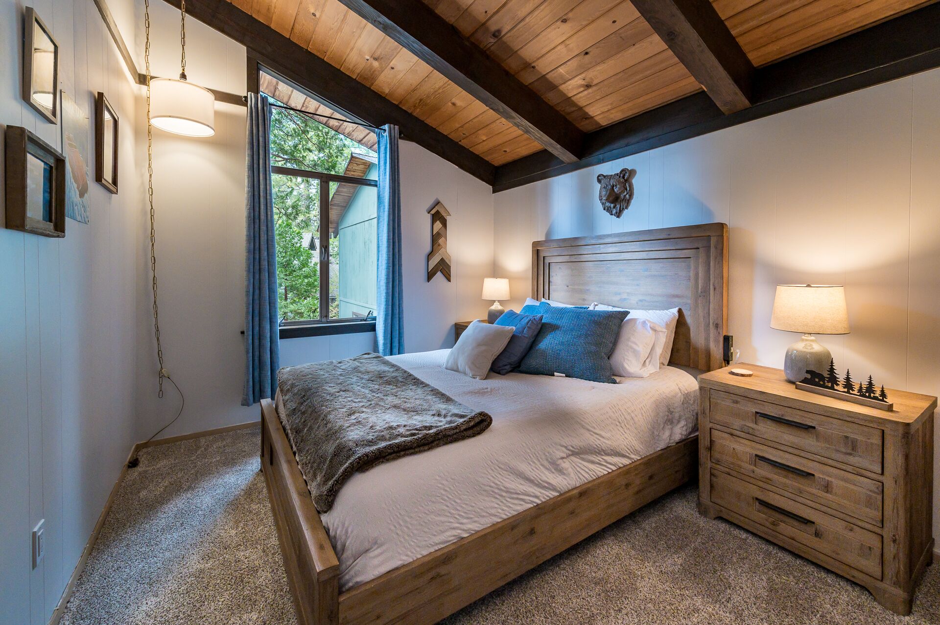 Un dormitorio con cama de madera, mesita de noche con lámpara y vistas a árboles por la ventana.