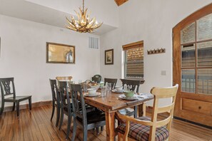 Une salle à manger avec une table en bois dressée pour quatre personnes, entourée de chaises, d’un lustre et d’œuvres d’art accrochées aux murs.