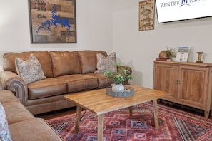 A living room with a brown leather sofa, a wooden coffee table, a patterned rug, and a cabinet with a sign on top.