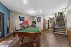 Une table de billard ornée d’un drapeau américain, une console de jeux vidéo et un tableau représentant un cerf, le tout dans un couloir au sol en bois et doté d’un escalier.