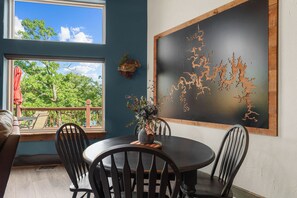 A dining area with a round table, four chairs, a vase with flowers, and a large abstract wooden art piece on the wall.