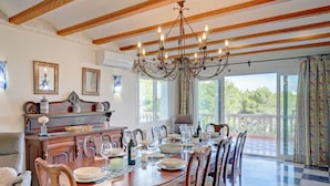 Een eetgedeelte met een ronde tafel gedekt voor zes personen, omringd door houten stoelen. Een kroonluchter hangt aan het plafond, en de kamer heeft een balkon met gordijnen.