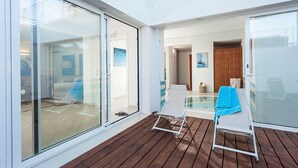 A modern room with a glass door leading to a balcony, a wooden deck, and a glimpse of a bedroom with a bed and a blue towel on a chair.