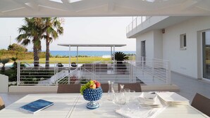 An outdoor dining area with a view of the sea, a white table with a book, a fruit bowl, and a pitcher.