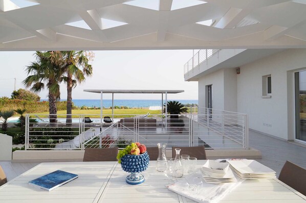 An outdoor dining area with a view of the sea, a white table with a book, a fruit bowl, and a pitcher. - Wonderful private villa with private pool, WIFI, sauna, hot tub, A/C, TV, patio and panoramic view (Marina Di Modica)