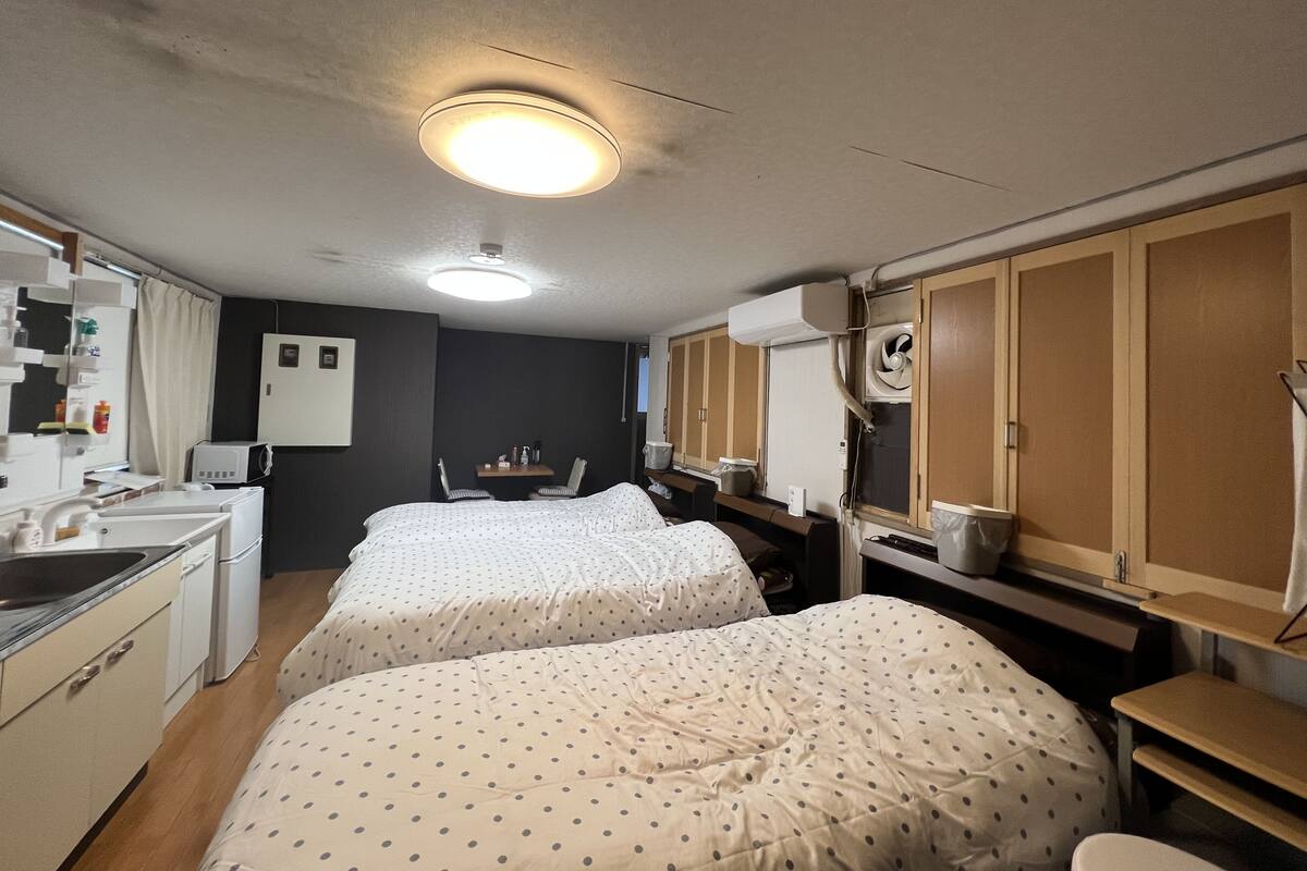A compact hotel room with two beds, a kitchenette, a small table, and a ceiling fan.