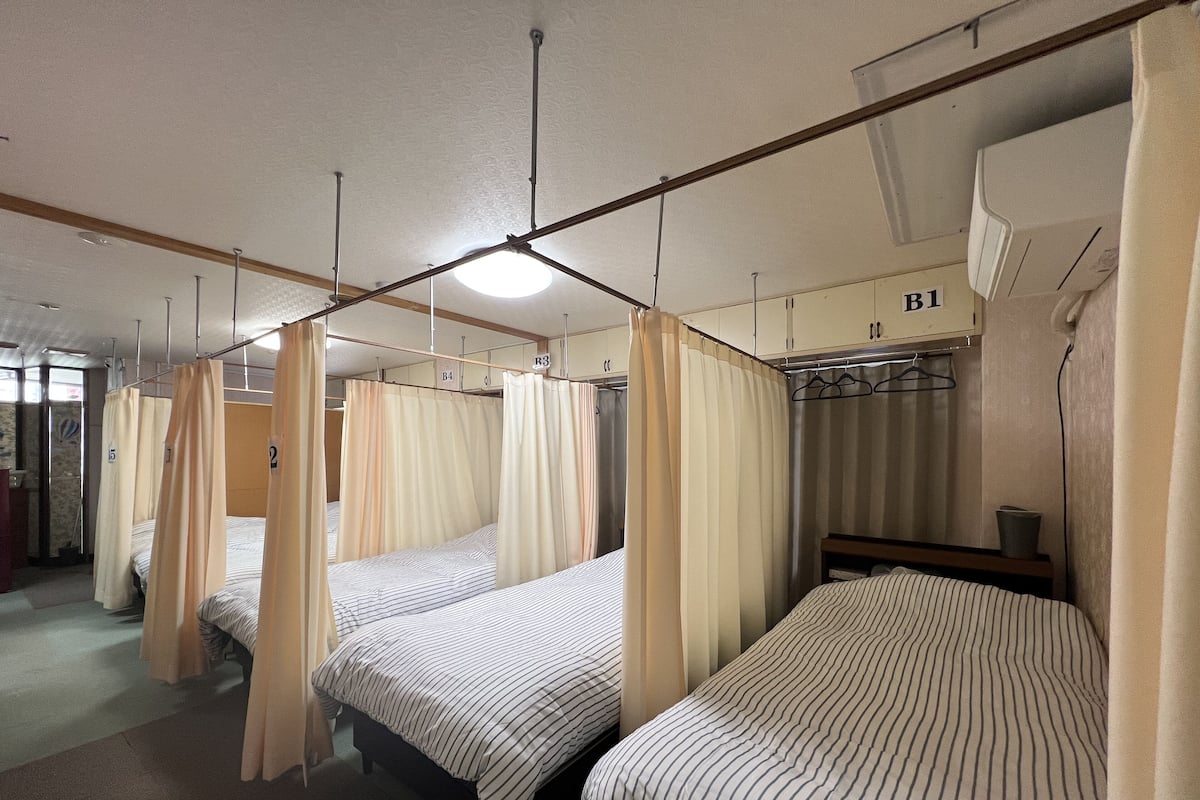 A hotel room with four single beds, each with striped bedding and a canopy. There is a wooden headboard, a bedside table, and a hanging light fixture.
