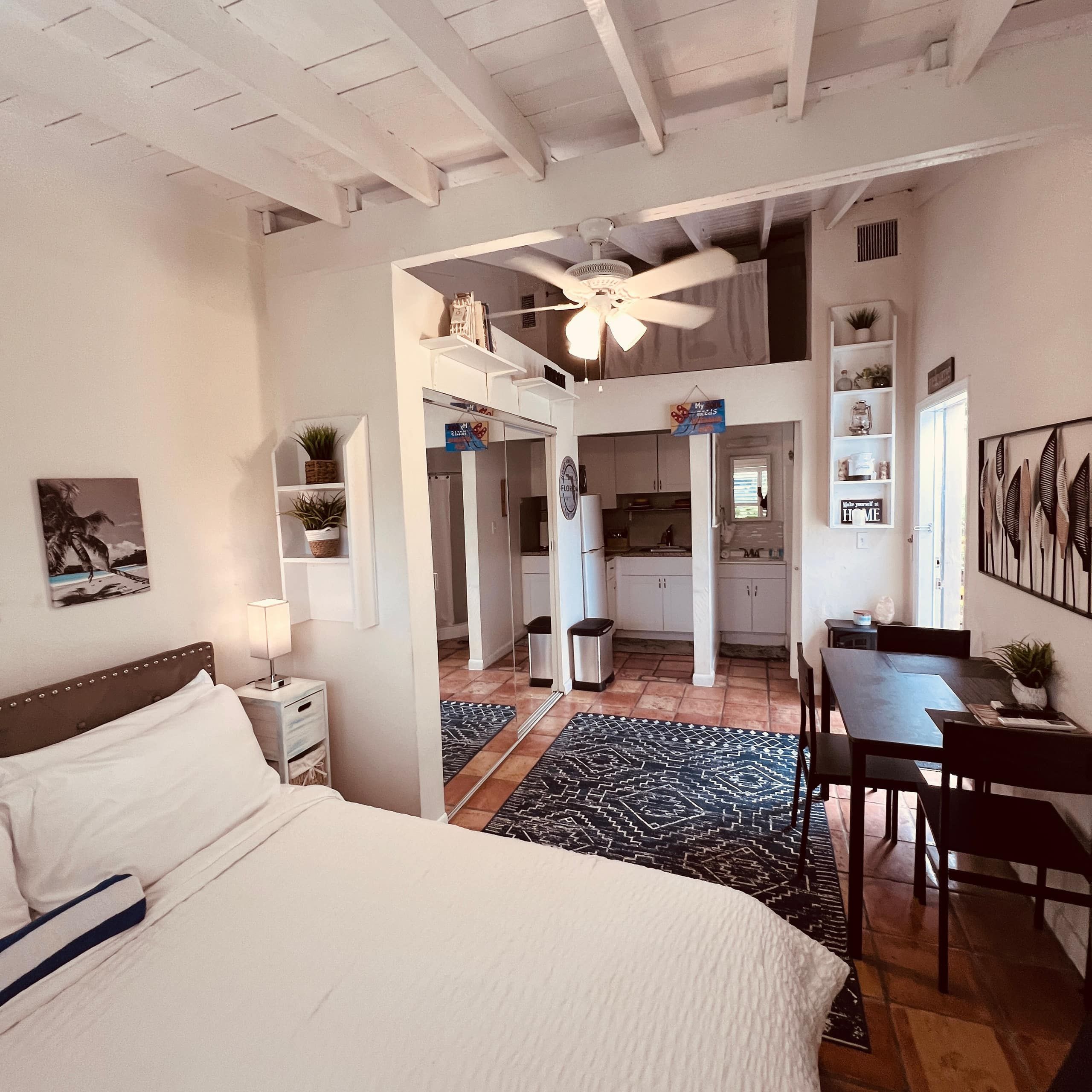 A bedroom with a bed, a desk, and a dining table. There is a kitchen in the background.