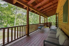 A covered porch with wooden railings, a cushioned seating area, and a view of a forested area.
