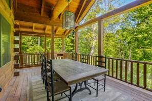 A wooden deck with a table and chairs, surrounded by a railing, overlooking a forested area.