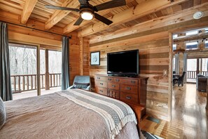 A wooden cabin bedroom with a bed, a dresser, a television, and a balcony with a view of trees.