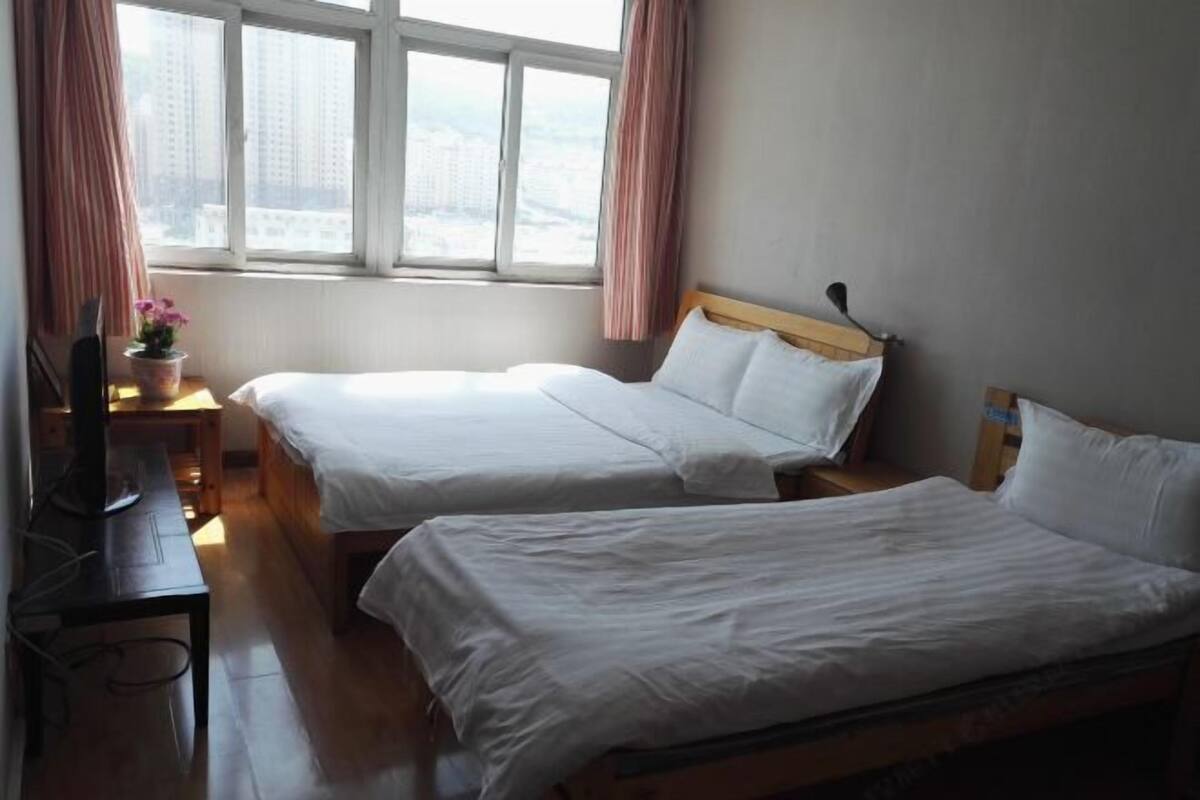 A hotel room with two beds, a desk with a computer, a window with curtains, and a view of buildings.