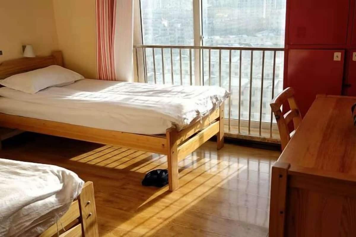 A room with a wooden bed, a small desk, and a window with a view of buildings.