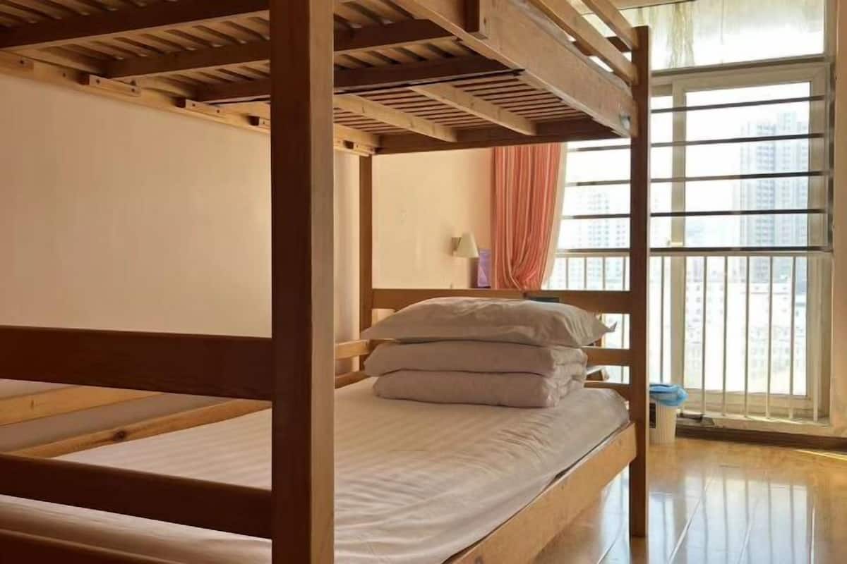 A bunk bed with a wooden frame, a white mattress, and a pillow. There is a window with a view of buildings outside.