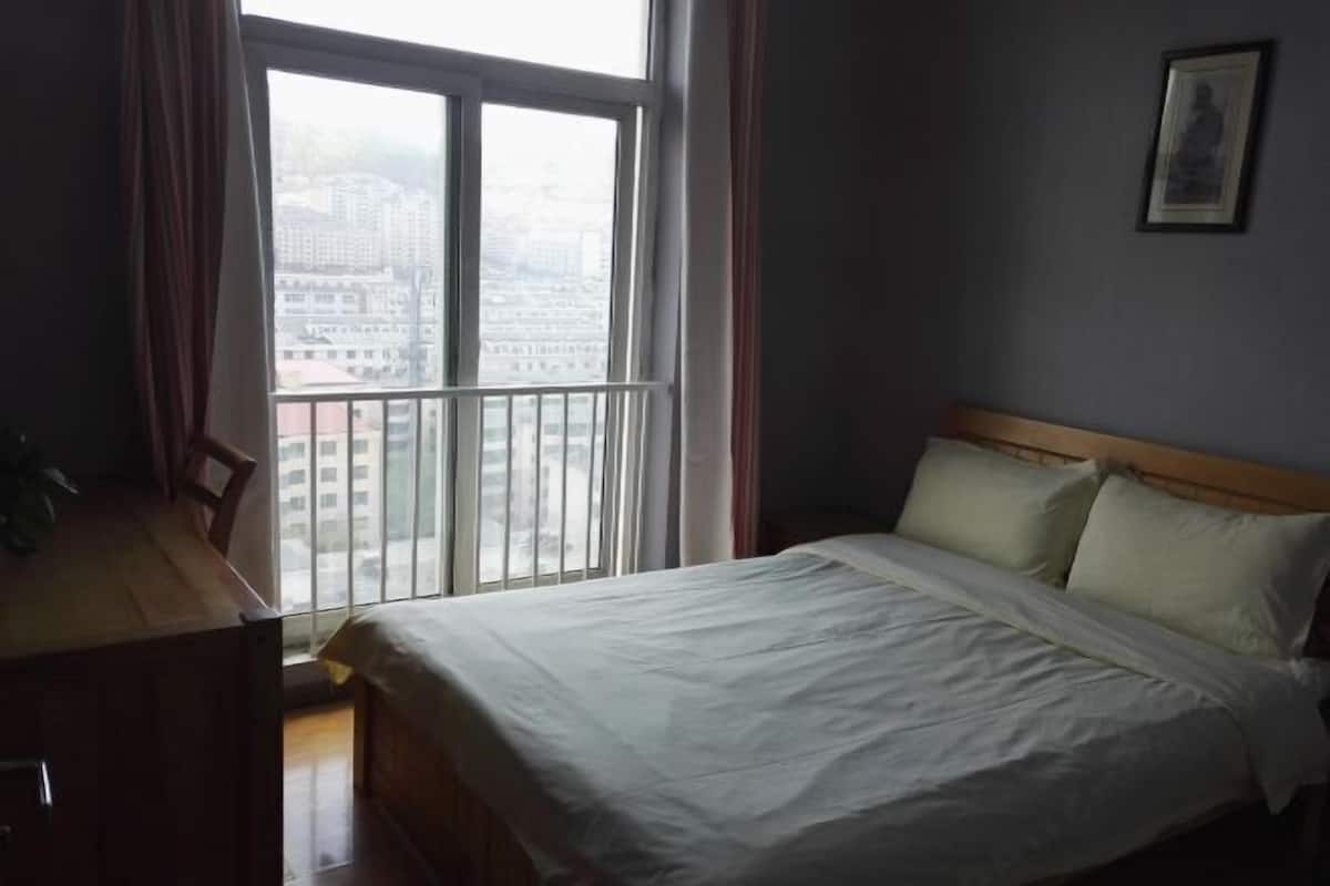A bedroom with a bed, a desk, and a window with a view of buildings.