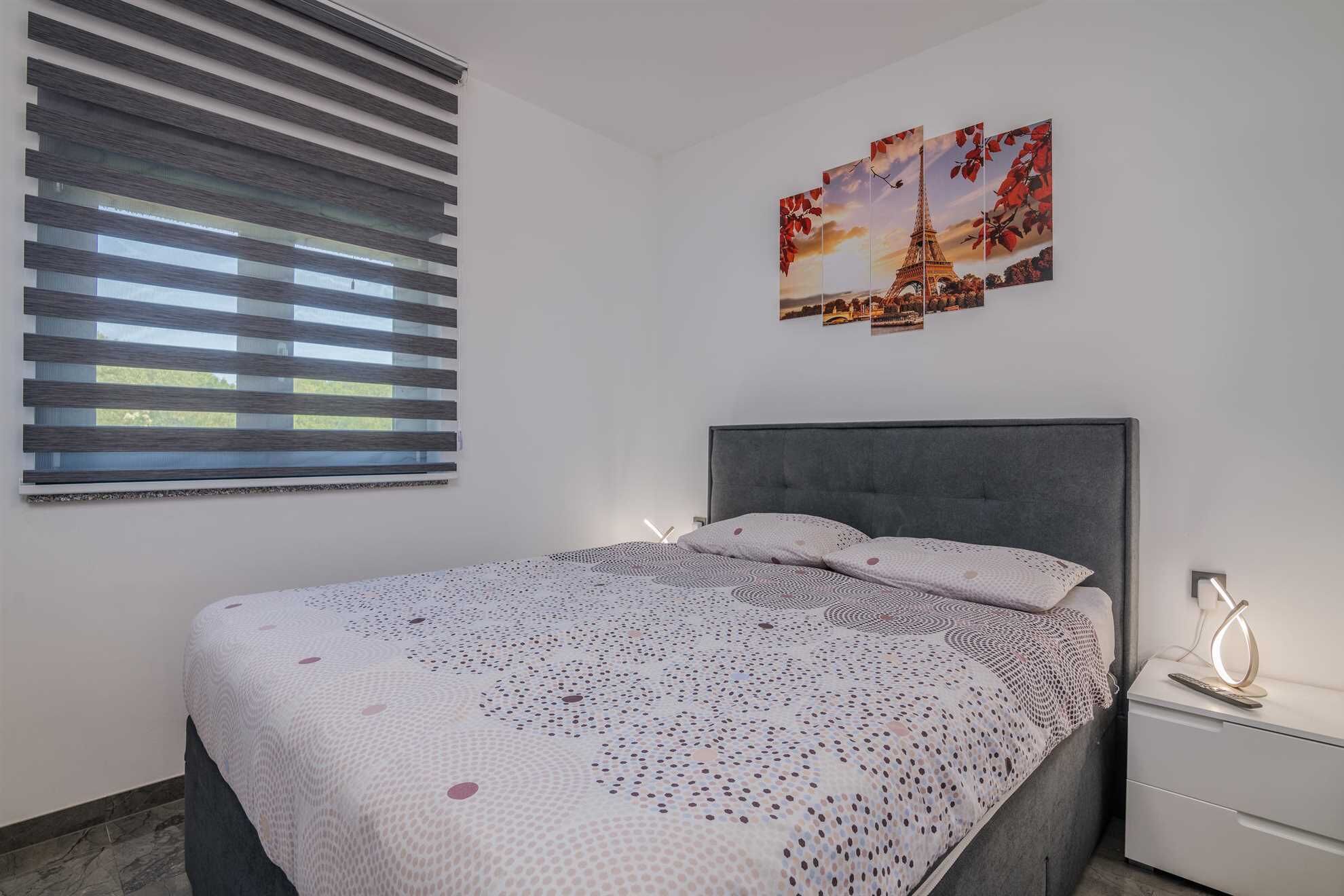 A bedroom with a bed, a nightstand, a wall-mounted picture of the Eiffel Tower, and a window with blinds.