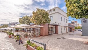 Une rue pavée avec des tables et des chaises à l’extérieur, un café et un bâtiment orné d’un grand arbre.