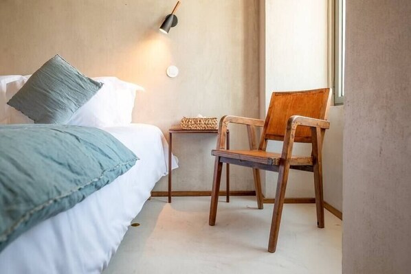 A bedroom with a bed, a wooden chair, a small table, and a wall-mounted lamp.