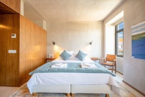A modern bedroom with a large bed, wooden wardrobe, bedside lamps, and a view of the city through a window.