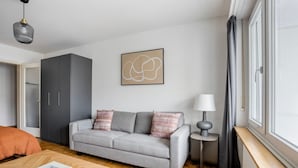 Un salon moderne avec un canapé gris, une petite table d’appoint avec une lampe, une table basse en bois et une grande fenêtre avec des rideaux.