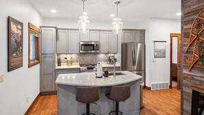 A modern kitchen with grey cabinets, stainless steel appliances, a marble island with bar stools, and pendant lighting. - The Dancing Bears Den (Whitefish)