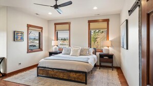 A bedroom with a bed, two nightstands, a ceiling fan, and large windows with blinds. - The Dancing Bears Den (Whitefish)
