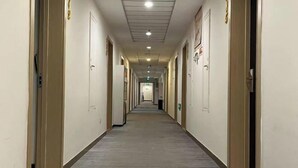 Un long couloir étroit, typique des hôtels, avec des portes des deux côtés, un sol carrelé et un plafond doté d’un éclairage encastré.