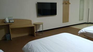 A hotel room with two beds, a TV mounted on the wall, a small round table, and a wooden desk with a mirror and a bottle of water.