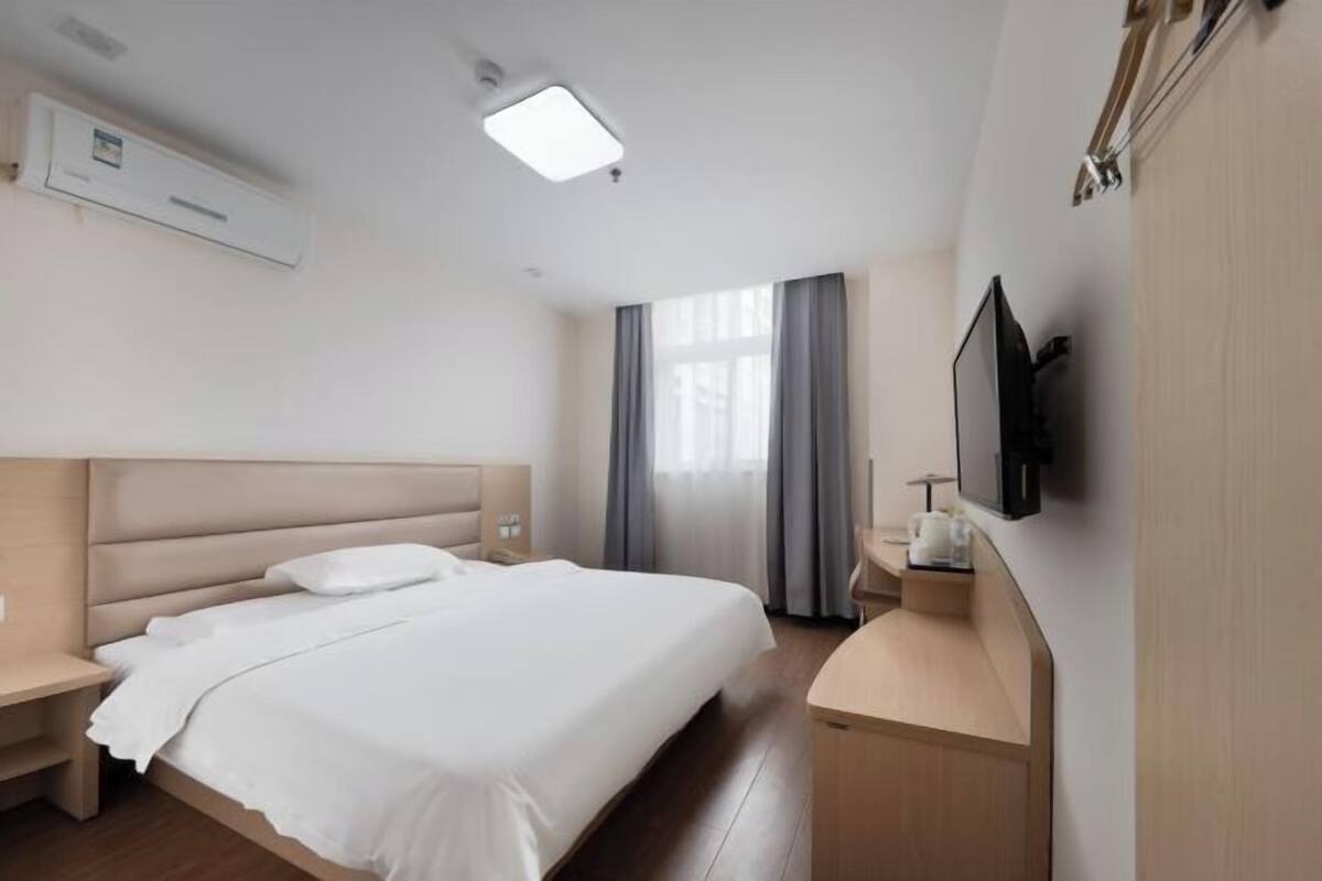 Hotellihuone, jossa on sänky, yöpöytä, televisio ja työpöytä tuolilla.